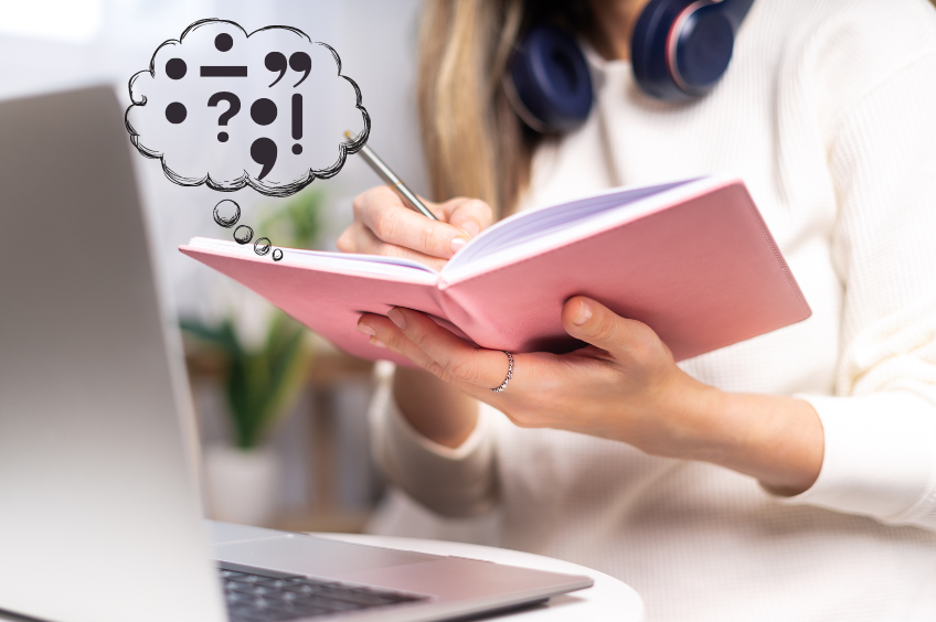 Woman sitting at her desk writing in a journal. She's surrounded by a thought bubble with various punctuation marks, including the semicolon
